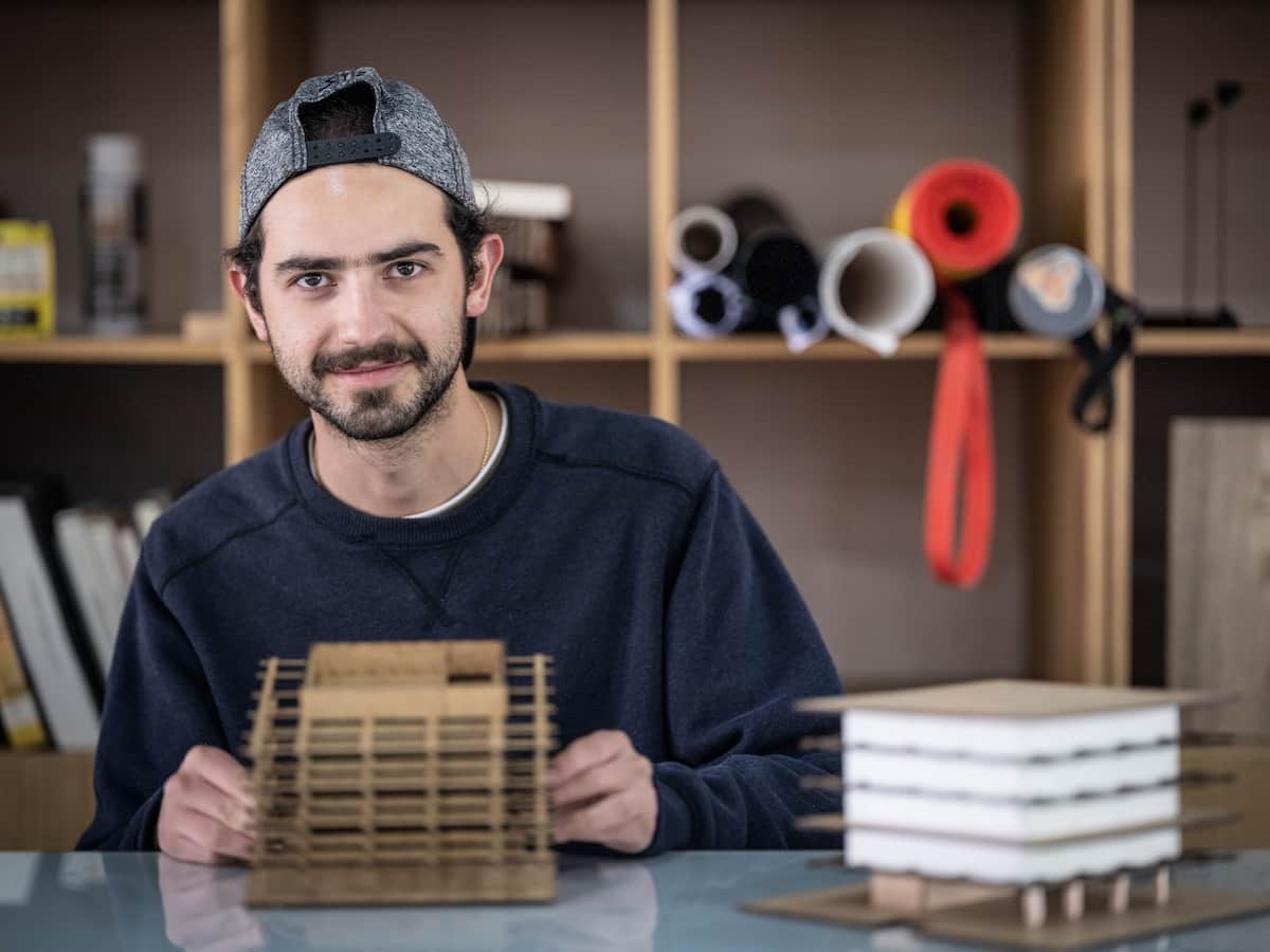 Estudiante de arquitectura mostrando maqueta de edificio de varios niveles frente a estantería con materiales.